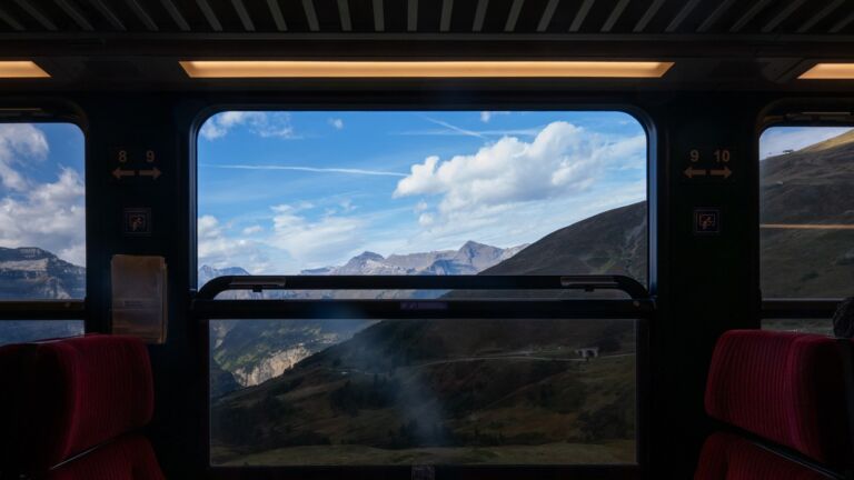 Blick durch ein Zugfenster auf eine Berglandschaft unter blauem Himmel – Bahnreise durch Europa